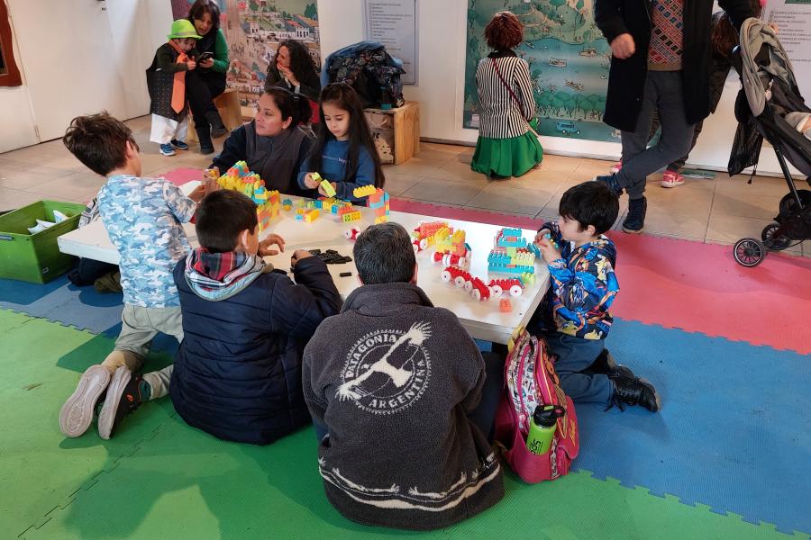 Grupo de personas, niños y adultos, en un espacio para infancias, sentados en el piso jugando con bloques de plástico.